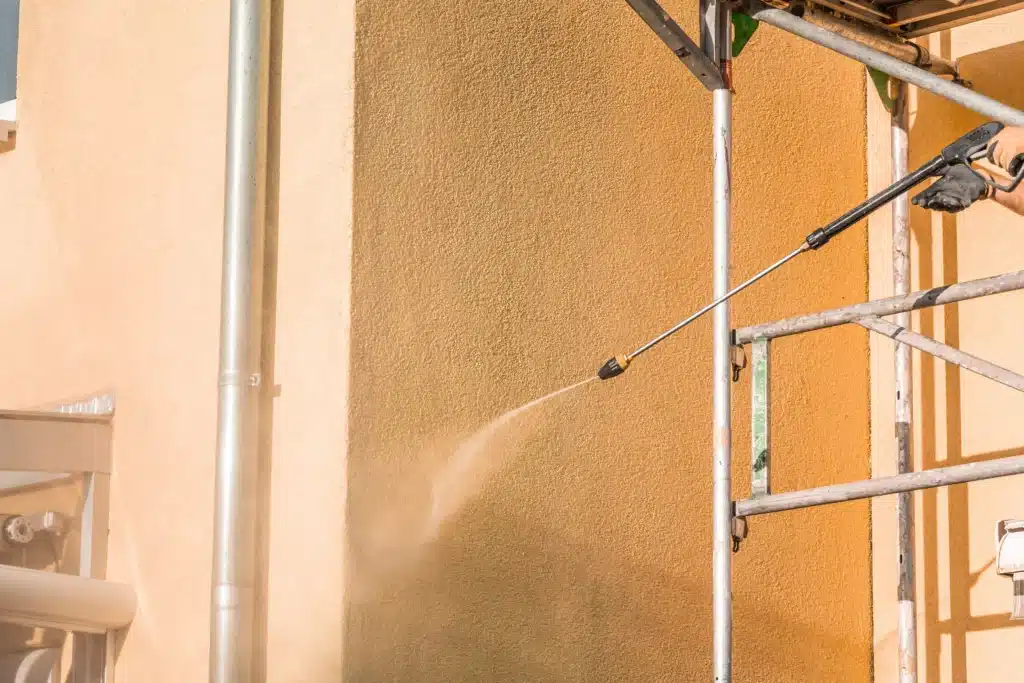 Main gantée utilisant un nettoyeur haute pression sur un mur en crépi. Le lavage révèle un mur orange vif près de l'échafaudage.