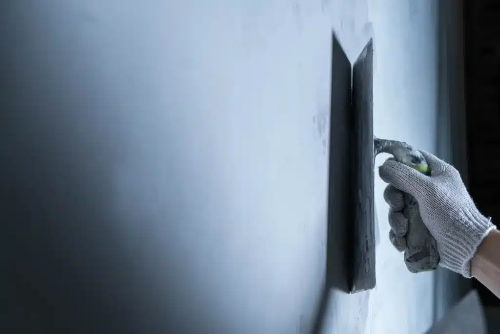Main gantée appliquant de l'enduit sur un mur avec un platoir de lissage. Travaux de crépissage et finition intérieure professionnels.