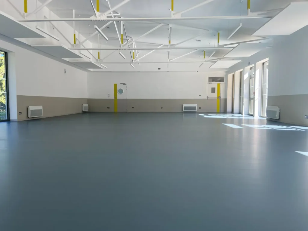 Grande salle vide moderne, sol gris uni et murs bicolores. Plafond design blanc avec poutres apparentes et accents jaunes vifs.