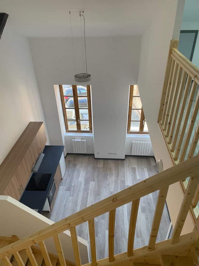 Duplex lumineux : cuisine aménagée et escalier bois moderne. Intérieur de loft lumineux. Balustrade bois d'escalier dominant une cuisine aménagée neuve et un salon avec deux grandes fenêtres.