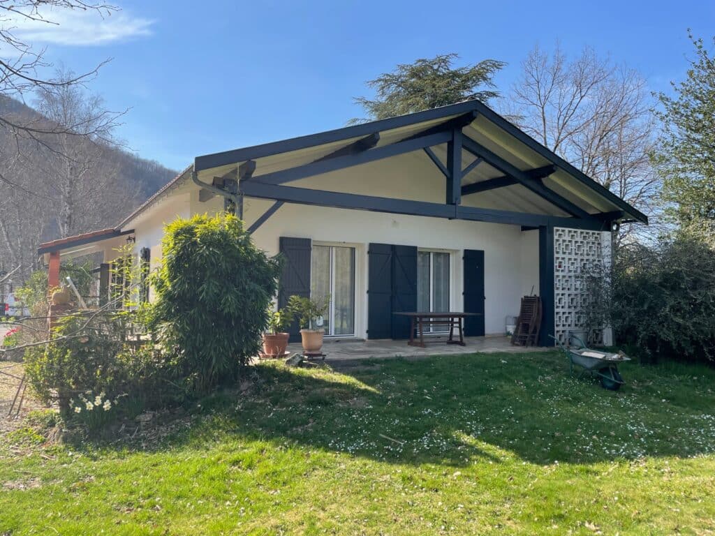 Maison blanche et terrasse couverte : design à ossature bois bleu foncé Maison blanche avec terrasse couverte par une ossature bois bleu foncé et volets assortis. Jardin fleuri devant la façade, avec montagne.