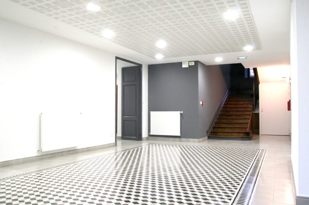 Hall rénové, carrelage ciment géométrique 3D noir/blanc. Mur gris, escalier bois sombre, radiateurs, plafond perforé lumineux.